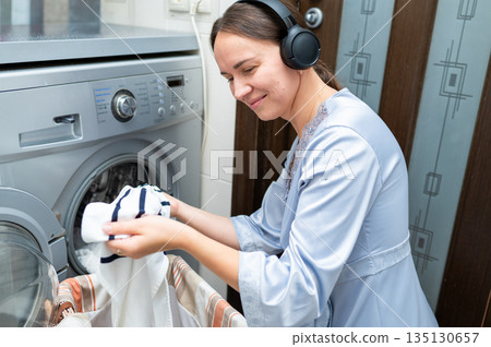 Cheerful housewife wearing headphones enjoying music while loading laundry into washing machine 135130657