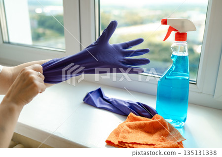 Detailed closeup of a womans hand putting on a glove by the windowsill with window cleaning spray and cloth ready for use 135131338