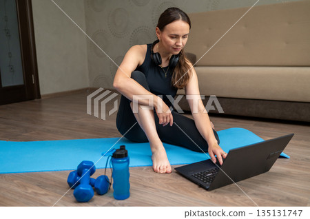 Woman sitting on yoga mat at home watching laptop before online pilates workout 135131747