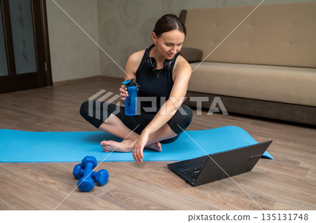 Woman holding water bottle on yoga mat at home watching laptop before pilates workout Woman holding water bottle on yoga mat at home watching laptop before pilates workout 135131748