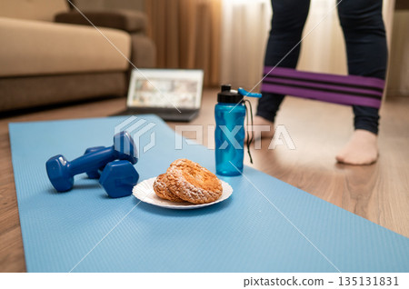 Dumbbells water bottle roller headphones and cookies on yoga mat with laptop and woman legs at home 135131831
