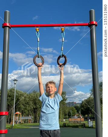 Teenage boy training on gymnastic rings at outdoor sports playground on a sunny summer day Teenage boy training on gymnastic rings at outdoor sports playground on a sunny summer day 135131960
