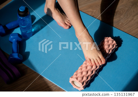 Female legs on foam roller doing myofascial massage with dumbbells and water bottle nearby 135131967