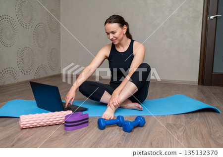 Young woman exercising at home with laptop on yoga mat using online fitness training program 135132370