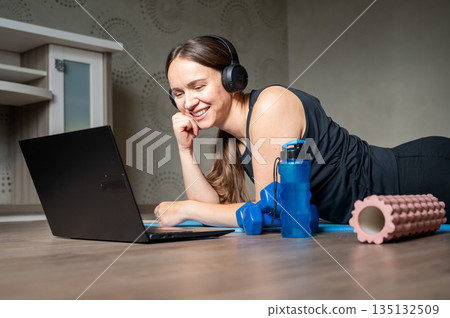 Happy woman lying on yoga mat with laptop headphones dumbbells water bottle and foam roller at home 135132509