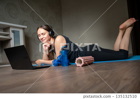 Woman lying on yoga mat with laptop headphones dumbbells water bottle and foam roller at home 135132510