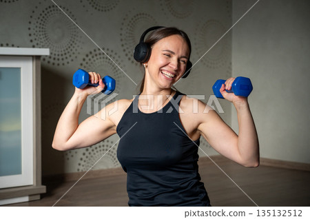 Smiling woman training with dumbbells at home wearing headphones 135132512