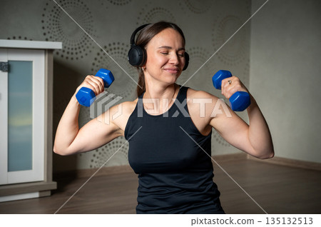 Smiling woman training with dumbbells at home wearing headphones 135132513