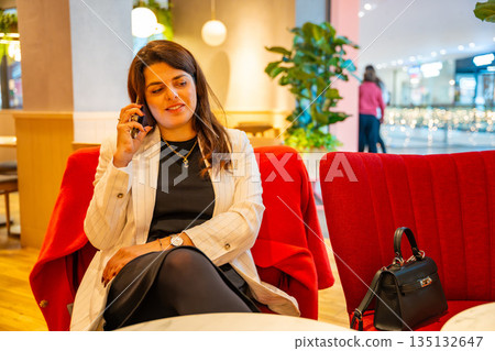 Woman sitting in cafe while waiting for order and talking on phone during shopping break. Everyday communication moment, leisure pause and modern urban lifestyle in real life commercial environment 135132647