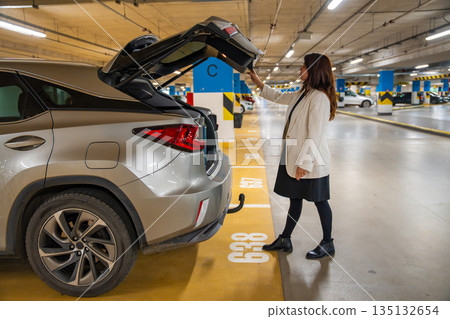 Woman closing car trunk by pressing button in public parking garage after shopping. Everyday departure routine, personal transport process and urban mobility moment in modern city environment Woman closing car trunk by pressing button in public parking garage after shopping. Everyday departure routine, personal transport process and urban mobility moment in modern city environment 135132654