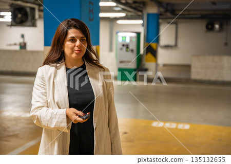 Woman unlocking car with remote control in public parking garage before leaving. Everyday departure moment, personal mobility and urban transport routine in modern European city environment 135132655