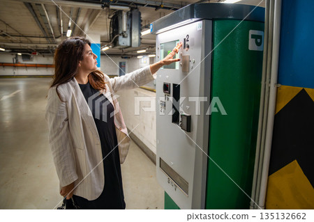 Woman paying for parking at automated payment machine in public parking garage. Everyday urban routine, personal transport management and mobility process in modern city environment Woman paying for parking at automated payment machine in public parking garage. Everyday urban routine, personal transport management and mobility process in modern city environment 135132662