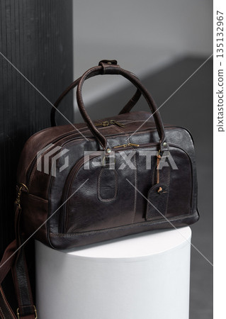 Luxury dark brown leather weekender travel bag on a white cylindrical pedestal in a minimalist modern studio with architectural textures. 135132967