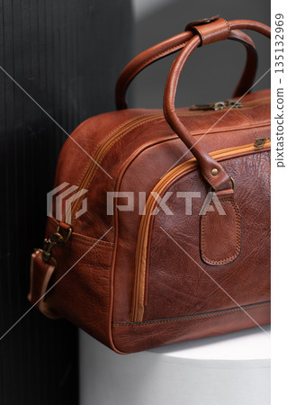 Luxury cognac leather weekender travel bag on a white cylindrical pedestal in a minimalist modern studio with architectural textures. Luxury cognac leather weekender travel bag on a white cylindrical pedestal in a minimalist modern studio with architectural textures. 135132969