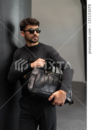 Confident man in sunglasses and charcoal knitwear holding a black leather briefcase in a modern minimalist interior. Confident man in sunglasses and charcoal knitwear holding a black leather briefcase in a modern minimalist interior. 135133079