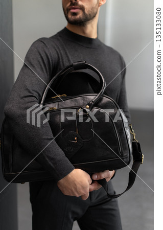 Confident man in charcoal knitwear holding a black leather briefcase while leaning against a textured pillar in a modern minimalist interior. Confident man in charcoal knitwear holding a black leather briefcase while leaning against a textured pillar in a modern minimalist interior. 135133080