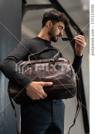 Confident man in charcoal knitwear holding a brown leather briefcase while leaning against a textured pillar in a modern minimalist interior. Confident man in charcoal knitwear holding a brown leather briefcase while leaning against a textured pillar in a modern minimalist interior. 135133086