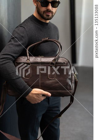 Confident man in sunglasses and charcoal knitwear holding a dark brown leather briefcase while leaning against a textured pillar in a modern minimalist interior. Confident man in sunglasses and charcoal knitwear holding a dark brown leather briefcase while leaning against a textured pillar in a modern minimalist interior. 135133088