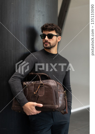 Confident man in sunglasses and charcoal knitwear holding a dark brown leather briefcase while leaning against a textured pillar in a modern minimalist interior. 135133090