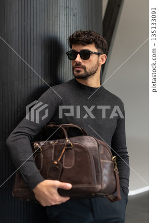 Confident man in sunglasses and charcoal knitwear holding a dark brown leather briefcase while leaning against a textured pillar in a modern minimalist interior. 135133091