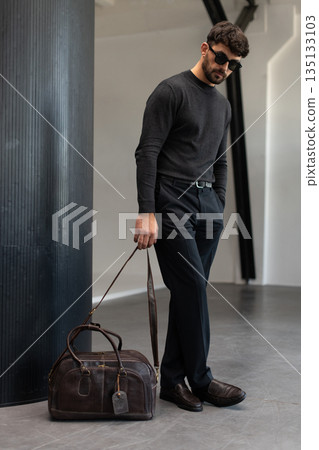 Confident man in sunglasses and charcoal knitwear holding a dark brown leather briefcase while leaning against a textured pillar in a modern minimalist interior. Confident man in sunglasses and charcoal knitwear holding a dark brown leather briefcase while leaning against a textured pillar in a modern minimalist interior. 135133103