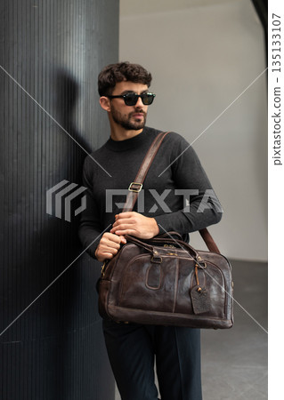 Confident man in sunglasses and charcoal knitwear holding a dark brown leather briefcase while leaning against a textured pillar in a modern minimalist interior. Confident man in sunglasses and charcoal knitwear holding a dark brown leather briefcase while leaning against a textured pillar in a modern minimalist interior. 135133107