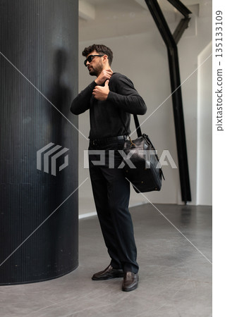 Confident man in sunglasses and charcoal knitwear holding a black leather briefcase while leaning against a textured pillar in a modern minimalist interior. 135133109