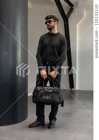 Confident man in sunglasses and charcoal knitwear holding a black leather briefcase while leaning against a textured pillar in a modern minimalist interior. Confident man in sunglasses and charcoal knitwear holding a black leather briefcase while leaning against a textured pillar in a modern minimalist interior. 135133110