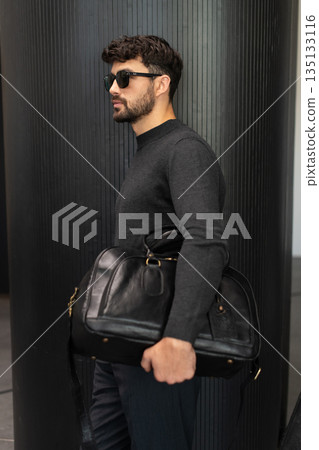 Confident man in sunglasses and charcoal knitwear holding a black leather briefcase while leaning against a textured pillar in a modern minimalist interior. Confident man in sunglasses and charcoal knitwear holding a black leather briefcase while leaning against a textured pillar in a modern minimalist interior. 135133116