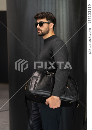 Confident man in sunglasses and charcoal knitwear holding a black leather briefcase while leaning against a textured pillar in a modern minimalist interior. 135133119