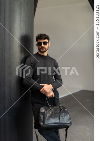 Confident man in sunglasses and charcoal knitwear holding a black leather briefcase while leaning against a textured pillar in a modern minimalist interior. 135133125