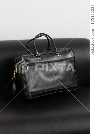 A professional black leather briefcase with dual handles and adjustable strap sits on a sleek black leather sofa in a minimalist studio setting. A professional black leather briefcase with dual handles and adjustable strap sits on a sleek black leather sofa in a minimalist studio setting. 135133222