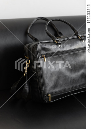 A professional black leather briefcase with dual handles and adjustable strap sits on a sleek black leather sofa in a minimalist studio setting. A professional black leather briefcase with dual handles and adjustable strap sits on a sleek black leather sofa in a minimalist studio setting. 135133243
