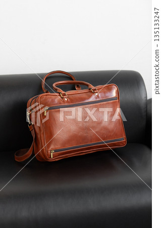A professional cognac leather briefcase with dual handles and adjustable strap sits on a sleek black leather sofa in a minimalist studio setting. 135133247