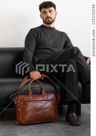 A professional male model in charcoal knitwear and tailored trousers sits on a black leather sofa holding a premium light brown leather briefcase. A professional male model in charcoal knitwear and tailored trousers sits on a black leather sofa holding a premium light brown leather briefcase. 135133248