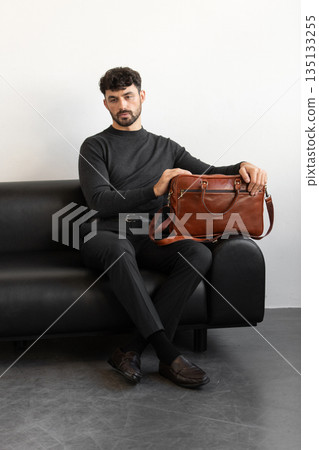 A professional male model in charcoal knitwear and tailored trousers sits on a black leather sofa holding a premium light brown leather briefcase. A professional male model in charcoal knitwear and tailored trousers sits on a black leather sofa holding a premium light brown leather briefcase. 135133255