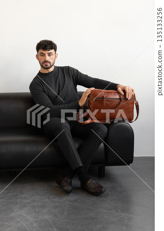 A professional male model in charcoal knitwear and tailored trousers sits on a black leather sofa holding a premium light brown leather briefcase. A professional male model in charcoal knitwear and tailored trousers sits on a black leather sofa holding a premium light brown leather briefcase. 135133256