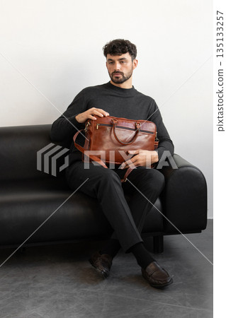 A professional male model in charcoal knitwear and tailored trousers sits on a black leather sofa holding a premium light brown leather briefcase. A professional male model in charcoal knitwear and tailored trousers sits on a black leather sofa holding a premium light brown leather briefcase. 135133257