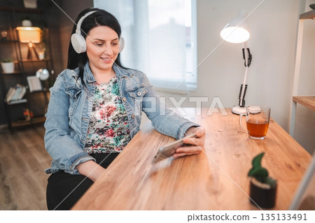 Woman enjoying music and using smartphone at home office Woman enjoying music and using smartphone at home office 135133491