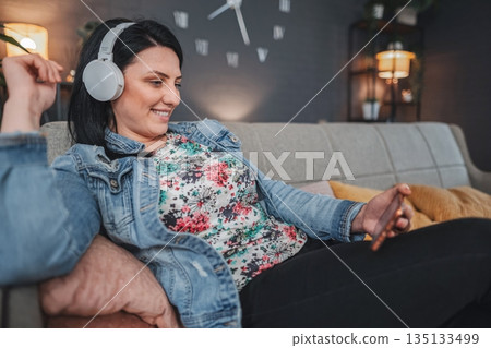 Woman relaxing on sofa listening music with headphones and using smartphone 135133499