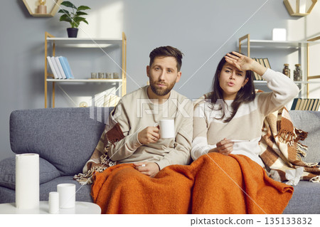 Sick young couple covered in blankets sitting on sofa at home with seasonal flu or cold. Sick young couple covered in blankets sitting on sofa at home with seasonal flu or cold. 135133832