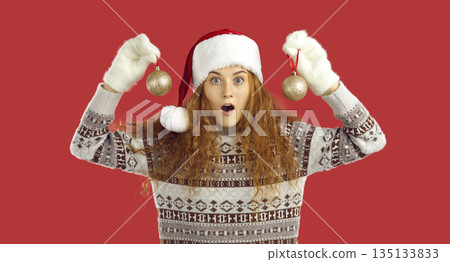 Portrait of astonished young woman with Christmas balls in hands on red background in studio. 135133833
