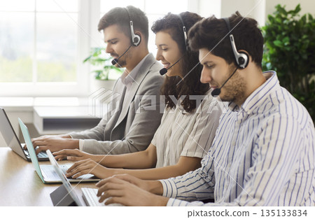 Call center agents with headsets work with laptops at desk, support customers Call center agents with headsets work with laptops at desk, support customers 135133834