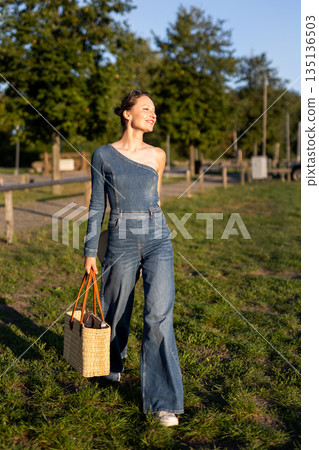 Young adult beautiful woman wear stylish jeans one-shoulder denim jumpsuit have fun walks outdoors warm sunny day holding straw bag hat hand. Female person carefree smiles enjoying countryside nature Young adult beautiful woman wear stylish jeans one-shoulder denim jumpsuit have fun walks outdoors warm sunny day holding straw bag hat hand. Female person carefree smiles enjoying countryside nature 135136503