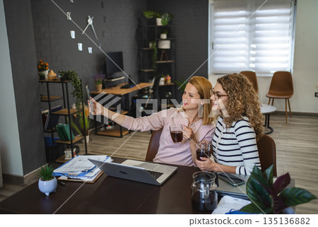 Two female colleagues are taking selfies and drinking coffee 135136882