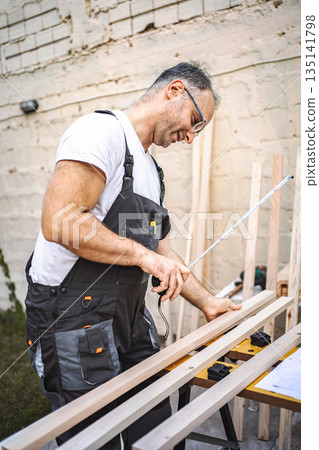 Mature male carpenter measuring wooden plank outdoors Mature male carpenter measuring wooden plank outdoors 135141798