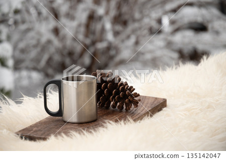 Metal mug with hot drink and pine cone on wooden board outdoors. Minimal winter composition with snowy background and copy space. 135142047