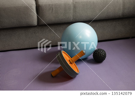 Home fitness equipment including exercise ball, ab roller and massage ball on yoga mat. Indoor workout and healthy lifestyle concept in cozy living room. Home fitness equipment including exercise ball, ab roller and massage ball on yoga mat. Indoor workout and healthy lifestyle concept in cozy living room. 135142079