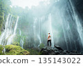 Woman stands on rocks admiring a majestic waterfall, Java, Indonesia. Tumpak Sewu 135143022