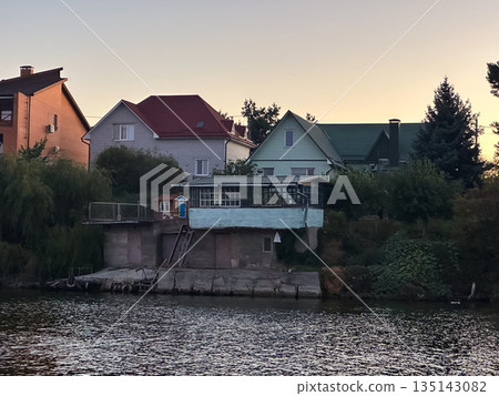 Houses by a riverbank at sunset with trees and calm water. Houses by a riverbank at sunset with trees and calm water. 135143082